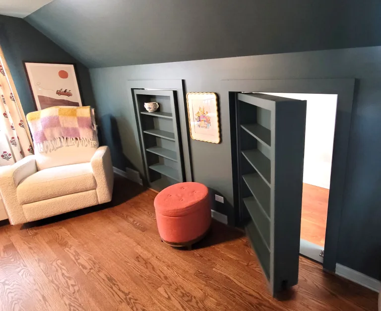 A custom hidden room design by Maya Construction featuring a secret doorway integrated into a dark grey floor-to-ceiling bookshelf next to a cozy seating area.